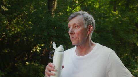 Sports and hydration concept. Active senior man drinking water outdoors, having break in sport activity in public park, tracking shot, slow motion