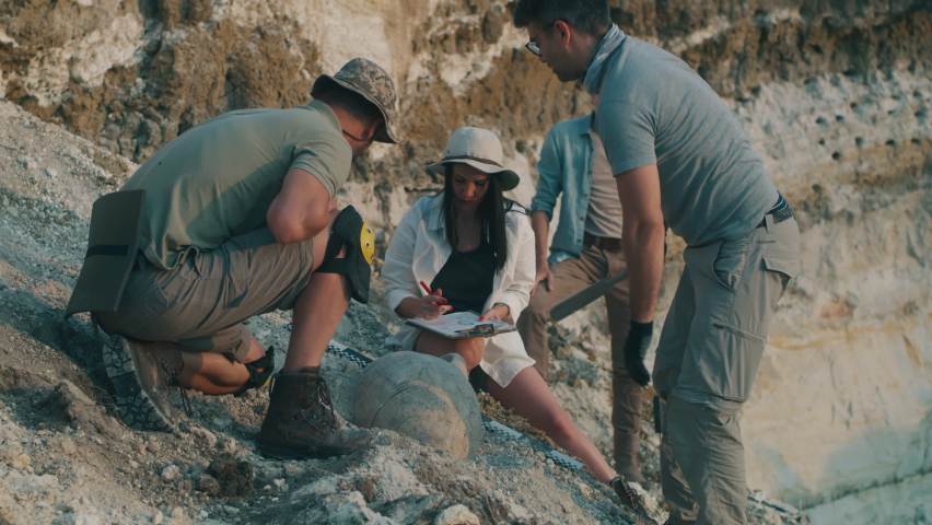 Men digging medieval vase from sand near male expedition leader supervising excavation and female colleague drawing sketches on archaeological site