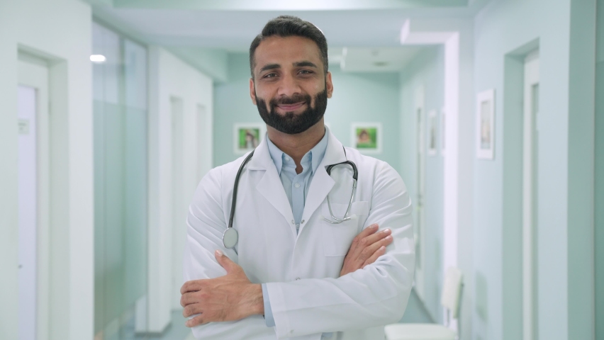 Smiling happy cheerful male Indian latin doctor medical worker in white robe with stethoscope around neck standing in modern hospital clinic with arms crossed looking at camera. Headshot portrait. Royalty-Free Stock Footage #1079079944