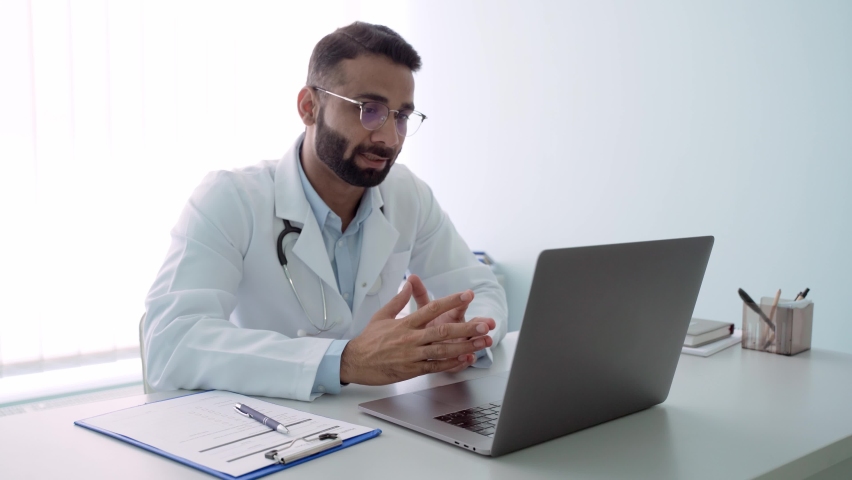 Indian male happy cheerful doctor therapist in modern clinic wearing eyeglasses and white coat uniform talking using laptop computer having videocall chat, consulting remotely. Telemedicine concept. | Shutterstock HD Video #1079079947
