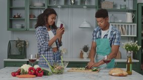 Millennial African American woman recording video on mobile phone of food blogger cooking fresh salad in kitchen. Man teaching to cook online.