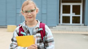 Portrait of pretty blonde schoolgirl standing in glasses with excercisebook in hand and backpack in front of sckool. 4K 60fps video