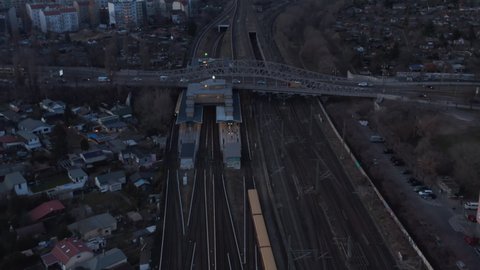 Aerial view of a passenger train passing by trees in Berlin, Germany moving towards station with people waiting under a bridge with cars moving surrounded by small hut houses during an early morning