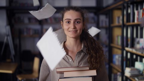Girl holding a lot of books in the library, paper sheets falling around her