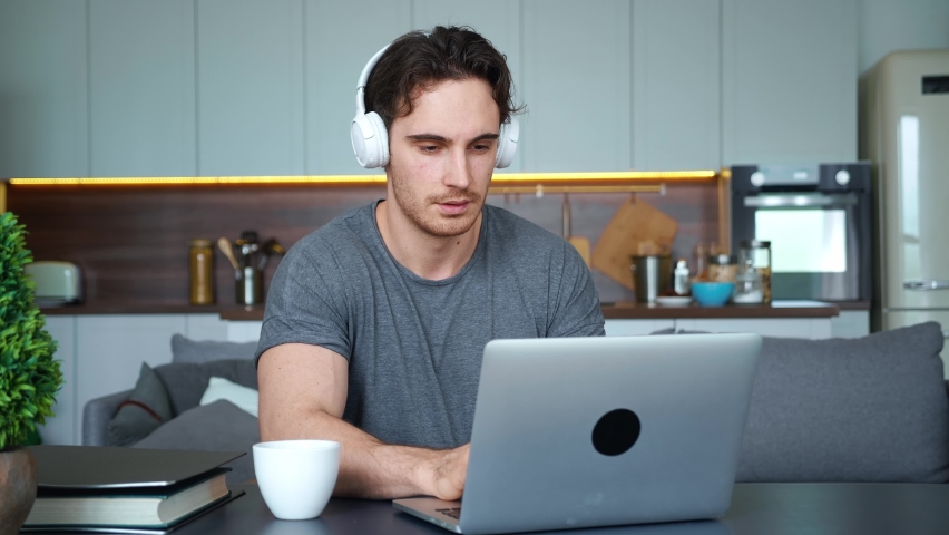 Handsome young man, attractive guy , freelance employee, businessman wearing wireless headphones, sitting at the table with cup of coffee and folder of documents, working on laptop remotely from home Royalty-Free Stock Footage #1079110280