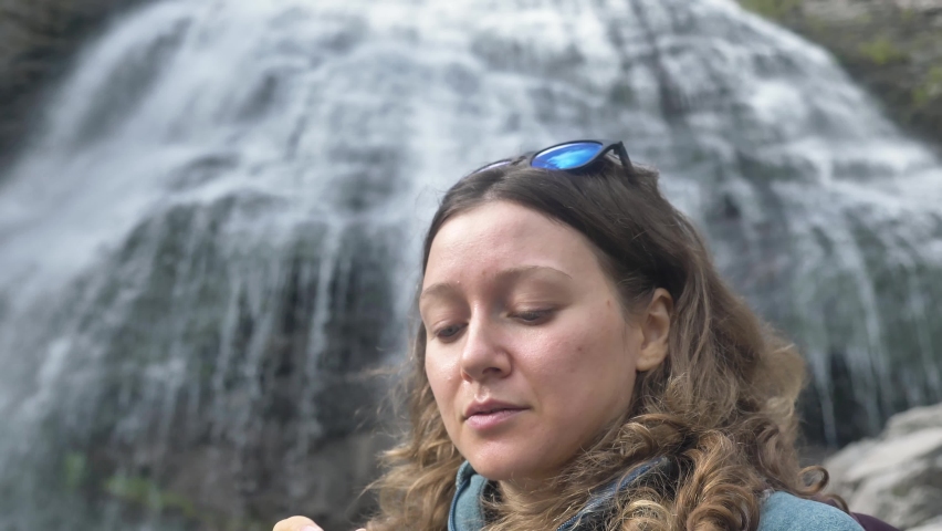 An active female tourist smears her face with sunscreen against the background of a large waterfall in the mountains in the open air, UV protection in the highlands