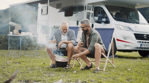 Mature man with senior father talking at campsite outdoors, barbecue on caravan holiday trip.