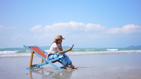 Asian woman resting on sunbed on the beach. Happy girl sitting on beach chair by the sea using smartphone for online shopping or text message. Female enjoy beach outdoor lifestyle on summer vacation