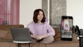 Blogger doing a live stream, sitting on a leather sofa with laptop. Talking in front of the camera on a stand and showing her clothes, in minimalist living room