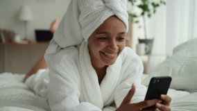Beauty blogger conducts stream. Spbd Cheerful African-American lady in white bathrobe uses videochat on mobile phone to speak with friends on bed in bedroom