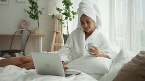 Beauty procedures relax at home. Spbd Pretty African-American lady in soft gown surfs internet social media on laptop holding cup on bed in stylish room