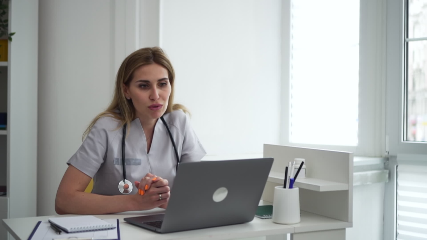 Female doctor working and speaking in front of laptop during online meeting in clinic office. Beautiful young woman talks with smile and looks at computer screen, gestures with hands and sits at Royalty-Free Stock Footage #1079118026