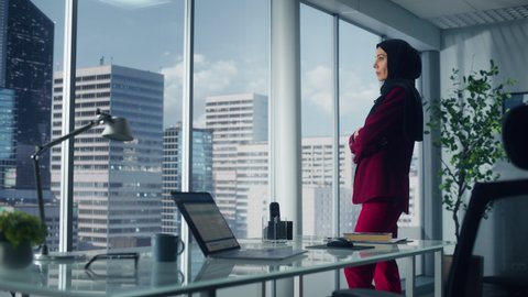 Successful Muslim Businesswoman Wearing Suit and Hijab Standing in Office Looking out of Window on Big City. Confident Female Digital Entrepreneur Planning Investment Strategy for e-Commerce Startup