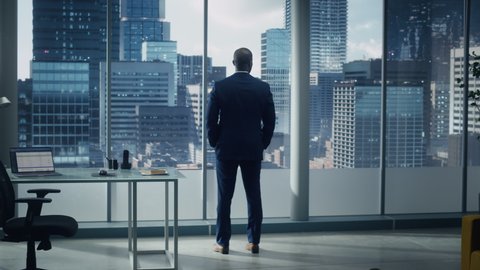 Thoughtful Black Businessman in a Tailored Suit Standing in Office Looking out of the Window on Big City. Successful Corporate Executive Thinking Investment Strategy for e-Commerce Startup. Back View