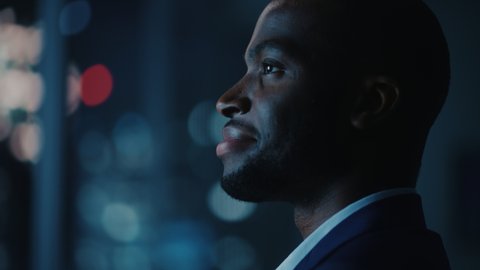 Night Office: Proud Close-up Portrait of Powerful Black Businessman Wearing Suit Standing, Looking out of the Window on a Big City. Ambitious African CEO Thinking of e-Commerce Investment Strategy