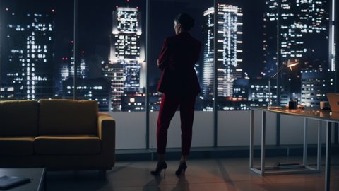 Night Office: Powerful Businesswoman Wearing Stylish Suit Standing and Looking out of the Window on a Big City. Female CEO Managing Environmental, Social and Corporate Governance for e-Business