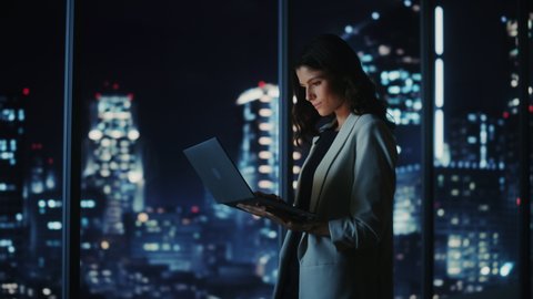 Big City Modern Office at Night: Successful Young Businesswoman Standing and Using Laptop. Beautiful Female Digital Entrepreneur Thinking of Investment Strategy for e-Commerce Project. Medium Shot