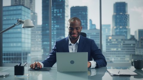 Portrait of Successful Black Businessman Working on Laptop Computer in His Big City Office. Smiling Digital Entrepreneur does Data Analysis for e-Commerce Strategy assessment. Zoom In Front View