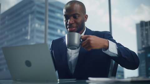 Portrait of Successful Black Businessman in Tailored Suit Working on Laptop Computer in His Big City Office. Digital Entrepreneur does Data Analysis, Cloud Computing for e-Commerce Strategy assessment