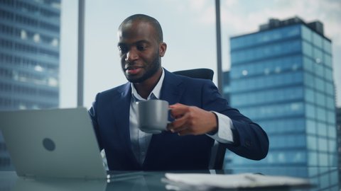 Successful Black Businessman in Tailored Suit Working on Laptop Computer on Top Floor Office Overlooking Big City. Successful Software Engineer does Data Analysis for His e-Commerce Startup Investment