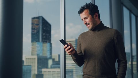 Thoughtful Arab Businessman Using Smartphone Standing in Office Looking out of the Window on Big City. Charismatic Digital Entrepreneur Plan Investment Strategy for e-Commerce Startup. Medium Arc Shot