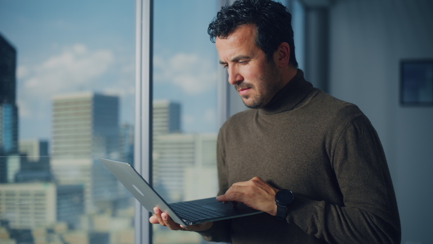 Professional Middle Eastern Businessman Using Laptop while Standing in Office Near Window on Big City. Successful Software Engineer Data Analysis for His e-Commerce Startup. Colorful Portrait