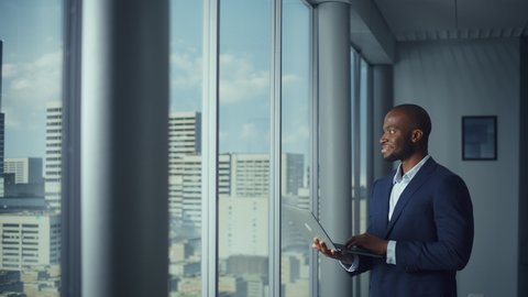 Thoughtful Black Businessman in Tailored Suit Using Laptop while Standing in Office Near Window on Big City. Successful Corporate CEO Doing Data Analysis management for e-Commerce Investment