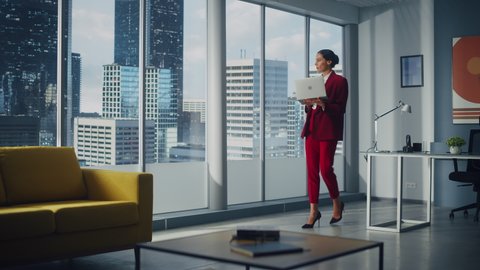Successful Caucasian Businesswoman Using Laptop While Standing in Office Looking out of Window on Big City. Confident Female Corporate CEO Managing Company Investment Strategy. Full Shot