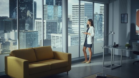Successful Caucasian Businesswoman Using Smartphone While Standing in Office Looking out of Window on Big City. Confident Female Digital Entrepreneur Developing e-Commerce Software Strategy. Full Shot