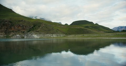 A beautiful view of the mountain lake at the foot of the densely vegetated hills. The tops of the rocky mountains are covered with thick clouds.