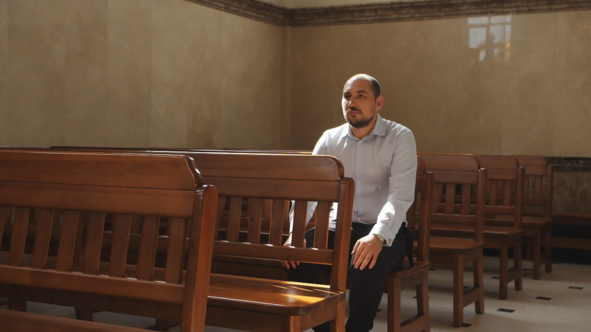 A man in a white shirt is sitting on a wooden bench, thinking and praying, looking around