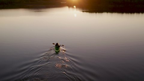 Fly away and up aerial FPV video of girl with red long hairs kayaking towards beautiful sunset, scenic Northern nature. Dark blue still water. Cinematic shot, sport vacation concept, Northern Sweden