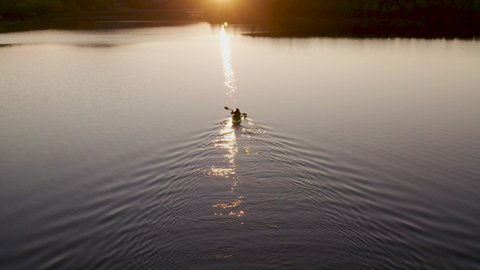 Following and approaching aerial FPV video of girl with red long hairs kayaking towards beautiful sunset, scenic Northern nature. Dark blue still water. Cinematic shot, sport vacation concept, Sweden