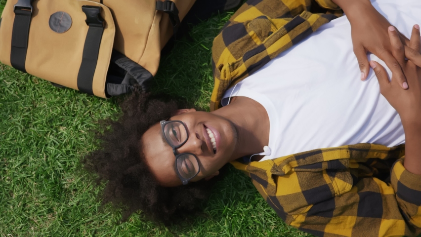 Top view of afro-american young man lying on green grass and looking at camera. Happy african male hipster in glasses relaxing on lawn in park and smiling at camera