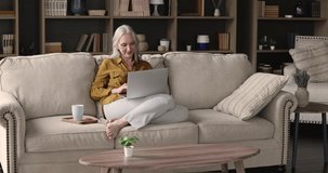 Beautiful aged 60s woman spend time alone at home relaxing on sofa with laptop and hot drink. Drinking coffee enjoy homey weekend with online fun, use modern wireless tech, internet connection concept