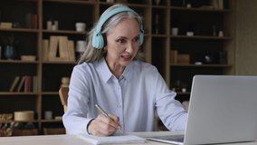 Older woman in headphones sit at desk in office, holds pen looks at copybook, discuss information with colleague or learner during business conference or on-line class. Virtual meeting event concept