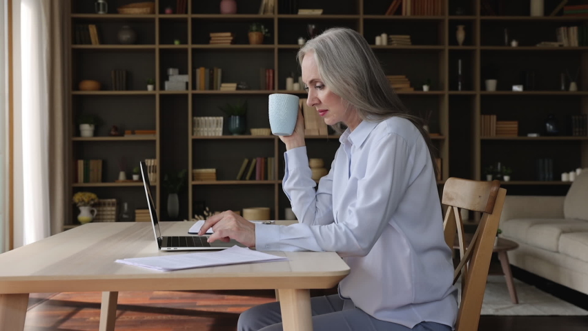 Serious mature woman sit at table in living room use laptop drink tea, spend time on internet, busy e-commerce shopping, learn new software, e services user, modern tech and older generation concept Royalty-Free Stock Footage #1079144432