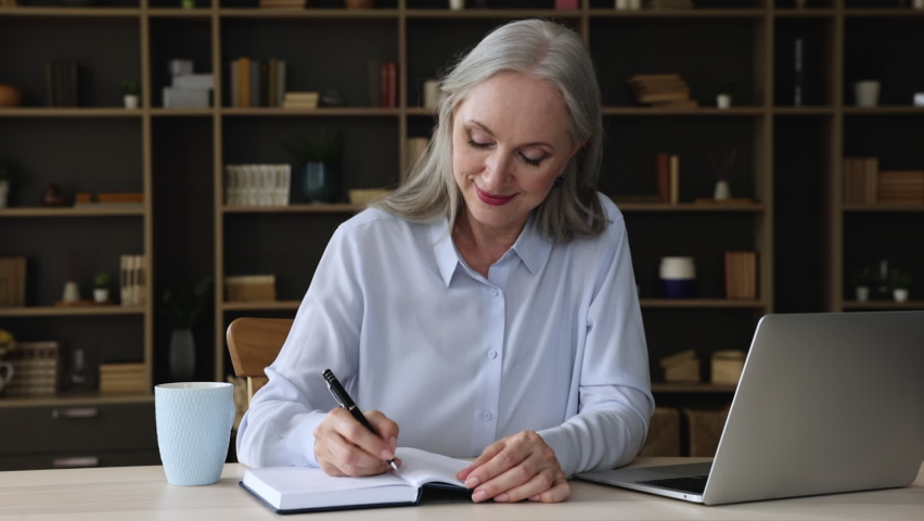Older grey haired businesswoman sit at workplace desk holds pen writing making notes in her organizer book, keep personal diary, fills it with useful information, create fresh business ideas and plans | Shutterstock HD Video #1079144456