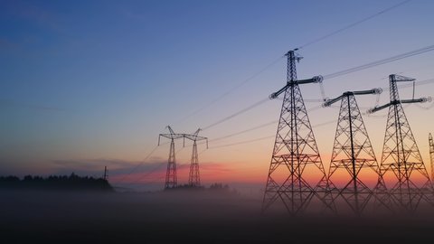 Aerial view high voltage steel power pylons in field covered with fog countryside. Misty early morning, dawn. Drone flight low over power transmission lines. Electric tower line, sunrise sunset