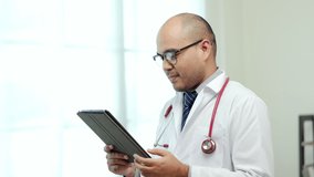 Asian doctor standing posing using tablet work from home. Positive male physician with stethoscope in hospital background. Healthcare and medical concept.