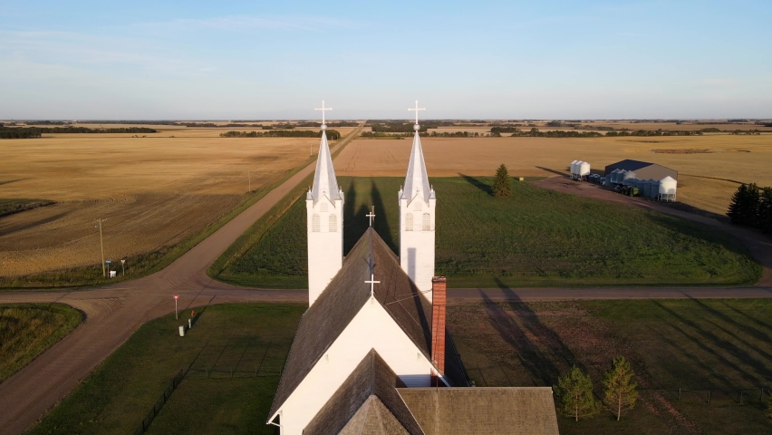 Aerial footage of St Peters roman catholic church in north American prairie during sunset. White building with blue roof on steeples.