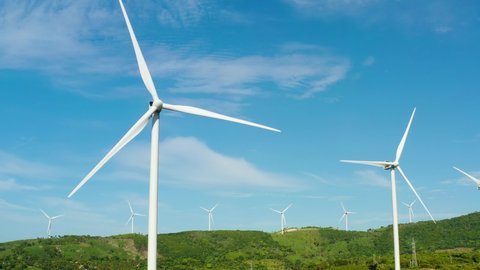 Beautiful white wind turbines are spinning large blades in a green field. Windmills in the mountainous area. Wind energy is modern green energy.