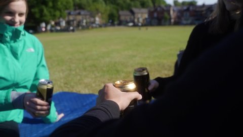 SHEFFIELD , South Yorkshire , United Kingdom (UK) - 08 18 2021: Slow-mo of three friends having a picnic and cheersing drinks in Endcliffe Park, Sheffield, England. Terraced houses, trees and neatly m