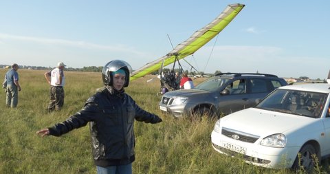 Berdsk, Siberia,  Russia, August 28.2021 Field airfield. A young woman in a helmet and a flight jacket smiles against the background of a hang glider with a motor .