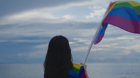Slow motion 4K. Person holding lgbtq flag in hand. Female waving rainbow LGBT flag on sunset blue sky with pride. Lesbian, Gay, Bisexual,  social movements. Freedom of love concept.