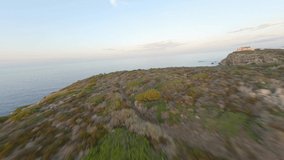 FPV video, view from above, stunning aerial view from an FPV drone flying at high speed over a rocky coastline with a lighthouse illuminated during a dramatic sunset. Faro di Capo Ferro, Sardinia.