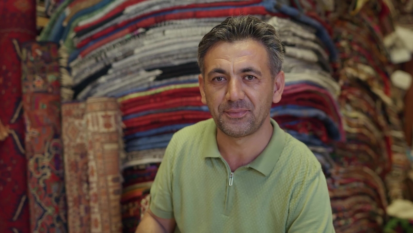 Turkish carpet seller in the carpet shop. The seller, sitting among the colorful authentic carpets, smiles at the camera. Traditional seller portrait.  | Shutterstock HD Video #1079184776