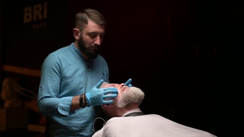 A master in a beauty salon rubs skin care cream into the face of an adult gray-haired man. Men's personal care salon