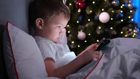 Small blond boy on Christmas night against background of Christmas tree, lying in bed with laptop in his hands and watching cartoons in dark room. Child plays games on laptop and learns remotely