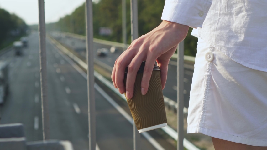 Female hand holds paper cup with coffee on  background of moving cars. Pedestrian city bridge near highway on sunny day. Coffee to go. Woman is holding drink while enjoying  view of passing cars.
